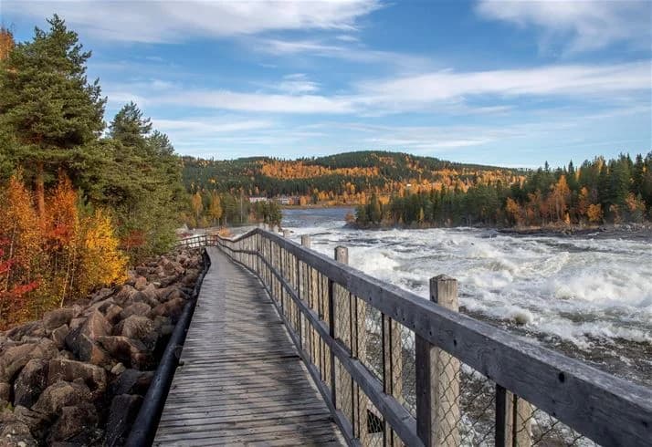 En pittoresk vy över en flod omgiven av klippor och strömmande vatten, med en bro som sträcker sig över floden. Moln pryder den blå himlen ovanför Storforsens camping.