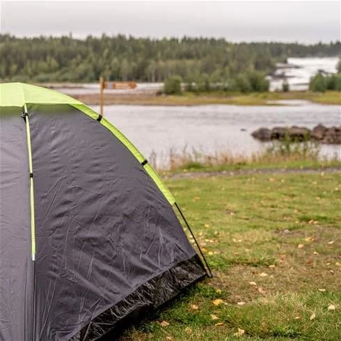 En tältplats vid Storforsens camping med flera tält som står utspridda i ett naturskönt parkliknande område. Utrustning för camping och vandring syns omkring, med en bakgrund av gröna skogar och berg.