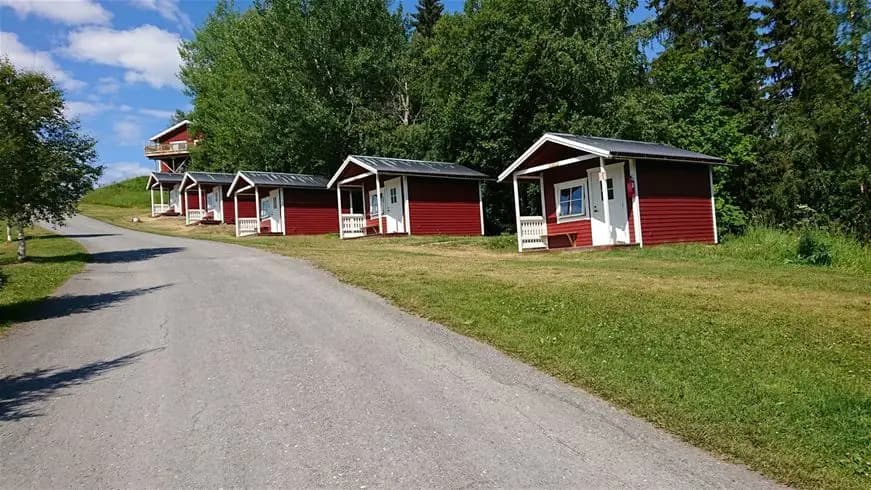 En grönskande campingplats med stuga vid Ristafallet, omgiven av öppna gräsytor och ett lantligt landskap.