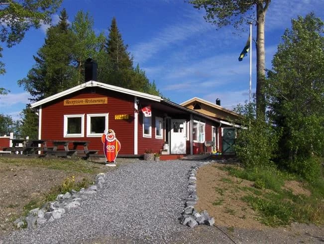 En campingplats med trädkantade områden där flera stugor med träpanel står utspridda. En uppfart leder in mot området, och det hela är omgiven av grönska.