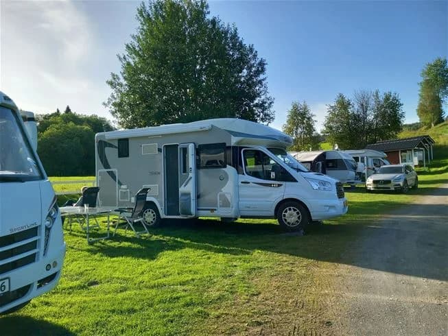 En husbil är parkerad vid Ristafallets camping, omgiven av grönska och en asfalterad väg, vilket ger en inbjudande och naturnära känsla.