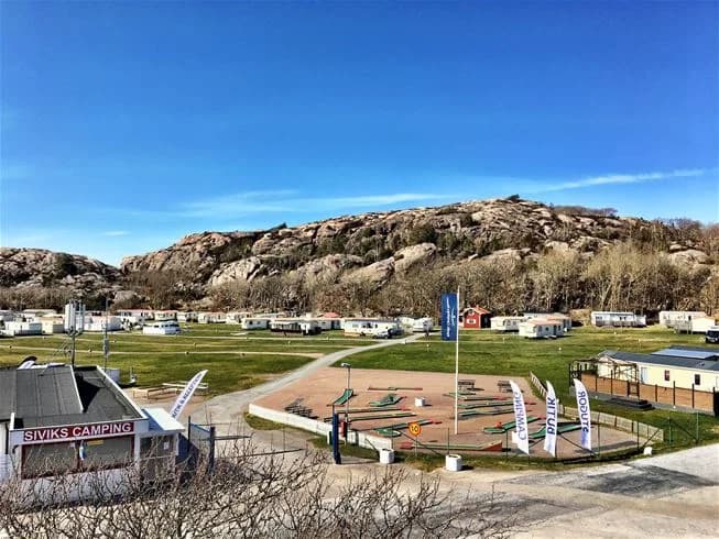 En naturskön campingplats med tält uppställda på en gräsbevuxen slätt, omgiven av skogsbeklädda kullar under en klar himmel. Perfekt för naturupplevelser och friluftsliv.