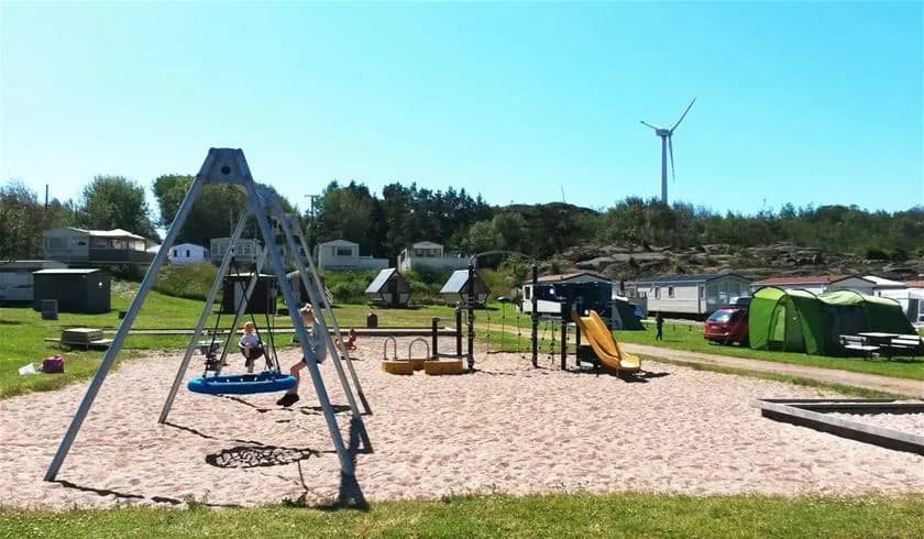 En lekplats med olika lekanordningar på Siviks Camping, omgiven av grönska och klarblå himmel, med en lätt vind som rör trädens löv.