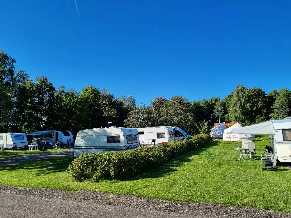 En bild av Tidaholms camping med flera husbilar och husvagnar parkerade på ett grusområde, omgivet av gröna träd, som utstrålar en trivsam och naturnära campingupplevelse.