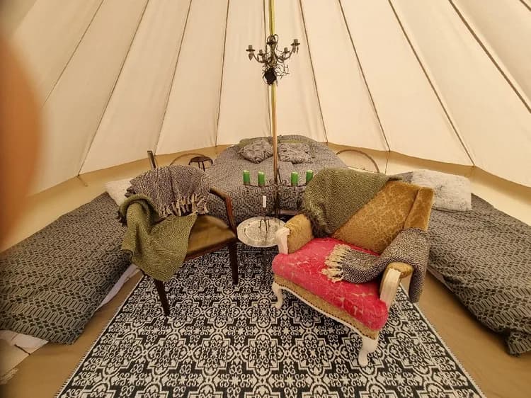Bild på insidan av en rymlig och stilfullt inredd yurt på Tidaholms camping, där möbler som sängar med linnelakan, ett bord, lampor och träinredning skapar en inbjudande miljö. Tältdukar och gardiner syns också, vilket förstärker känslan av bekvämt campingliv i naturen.