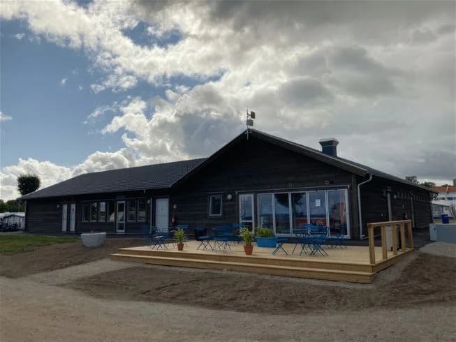 En liten stuga med rött tak och träfasad står på Grönhag Camping under en molnig himmel. Stugan har en vit dörr och är omgiven av grönska med en skuggig veranda.