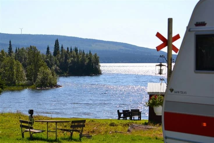 En fridfull sjö omgiven av tät barrskog, med tallar och granar dominerande landskapet. På vattnet flyter en ensam båt, och i bakgrunden syns böljande kullar. Scenen fångar den rustika och natursköna atmosfären vid Åkersjöstrand Camping.
