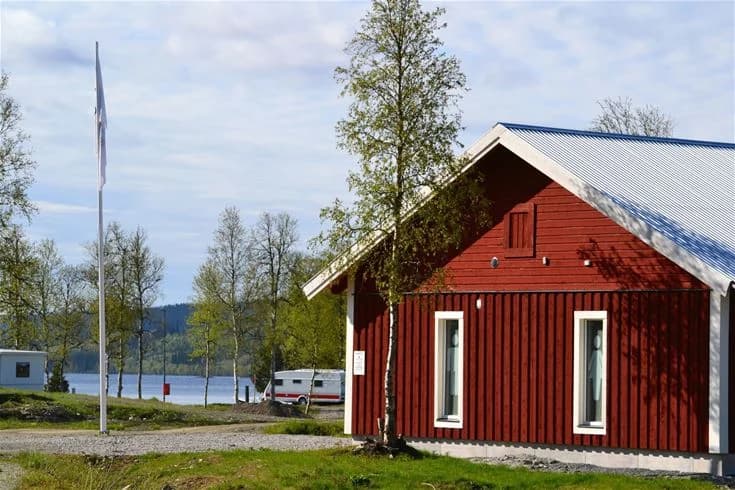 En idyllisk vy över Åkersjöstrand Camping med stugor och timmerstugor omgivna av tallar, beläget vid en sjö i ett pittoreskt landskap.