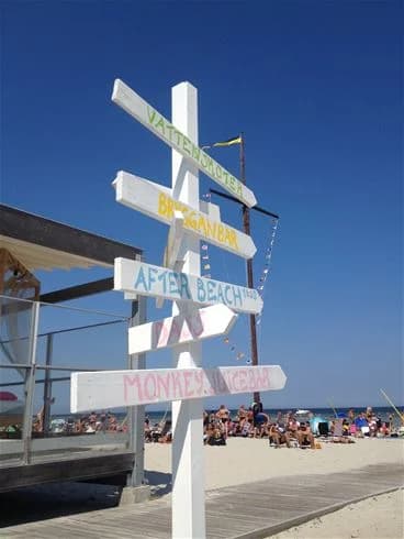 En solig dag på Köpingbaden camping med en sandstrand i förgrunden, campingstugor och flaggstänger synliga i bakgrunden, semesterfirare njuter av vinden och semestern.
