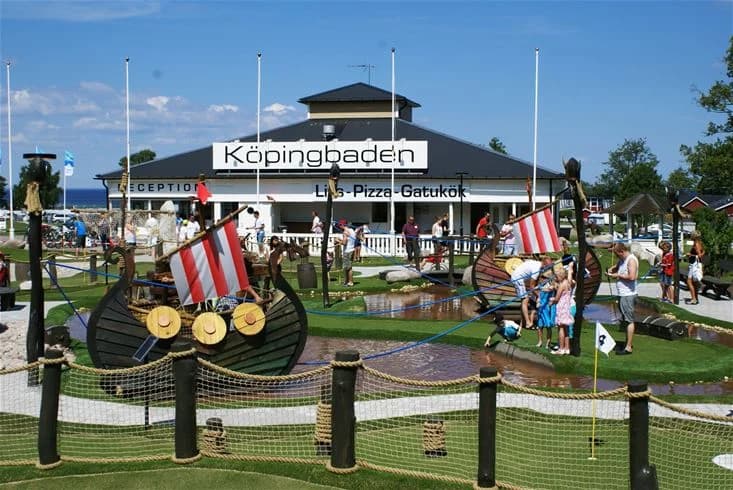 En solig dag på Köpingbaden Camping med grönskande gräsmatta och skuggiga områden. Flera flaggor vajar på höga stolpar och campingens öppna ytor välkomnar besökare för rekreation och avkoppling. En luftig baldakin ger extra skugga.