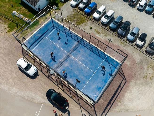 En padeltennisbana med grönt underlag syns inom ett inhägnat område av kedjelänkstaket på Köpingbaden Camping, en plats för rekreation och sportaktiviteter.