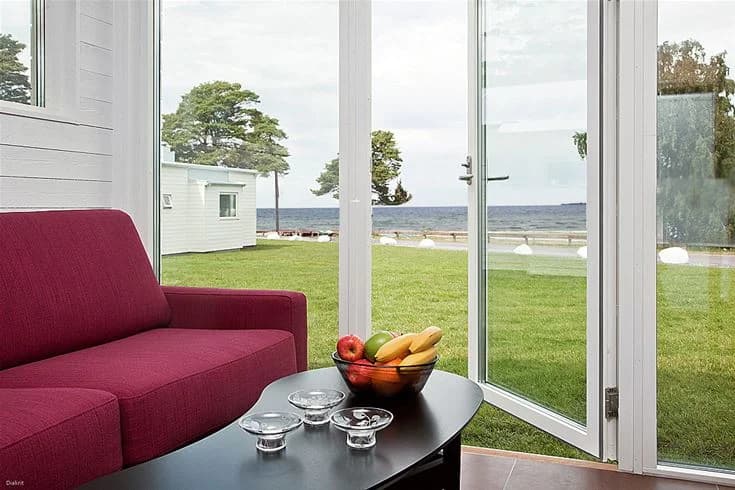 En campingstuga med en glasdörr och en koffeebord i skuggan på uteplatsen, omgiven av krukväxter och utemöbler under dagsljus.