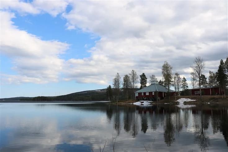 En vintrig landskapsbild vid Trehörningsjö Camping & Stugor, med snötäckta barrträd i förgrunden och en stilla sjö i bakgrunden, inbäddad i kvällsljusets dämpade sken.