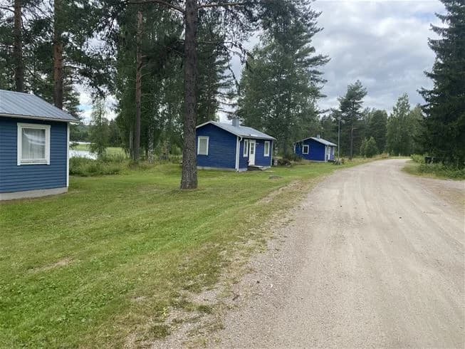 En öppen gräsyta vid Trehörningsjö Camping & Stugor, omgiven av ett lantligt landskap med små stugor och trädgårdsbyggnader i bakgrunden.