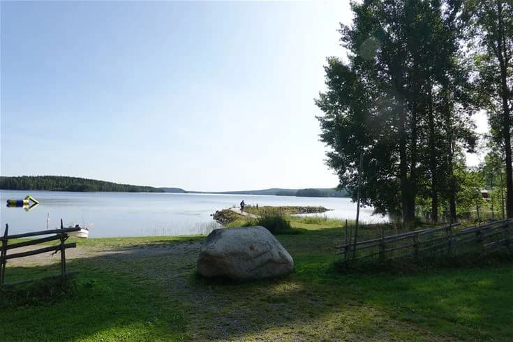 En somrig vy över en vacker sjö omgiven av grönska vid Trehörningsjö Camping & Stugor, med en bänk vid strandkanten och klart vatten i förgrunden.