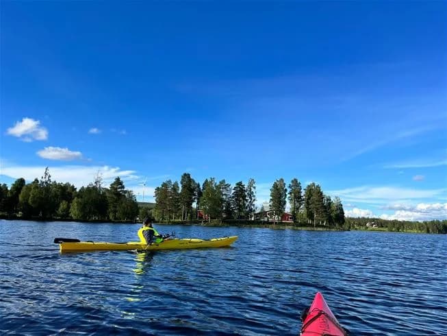 En person paddlar kajak på en lugn vattendrag nära trehörningsjö camping & stugor, omgiven av grönska och sommarlandskap.
