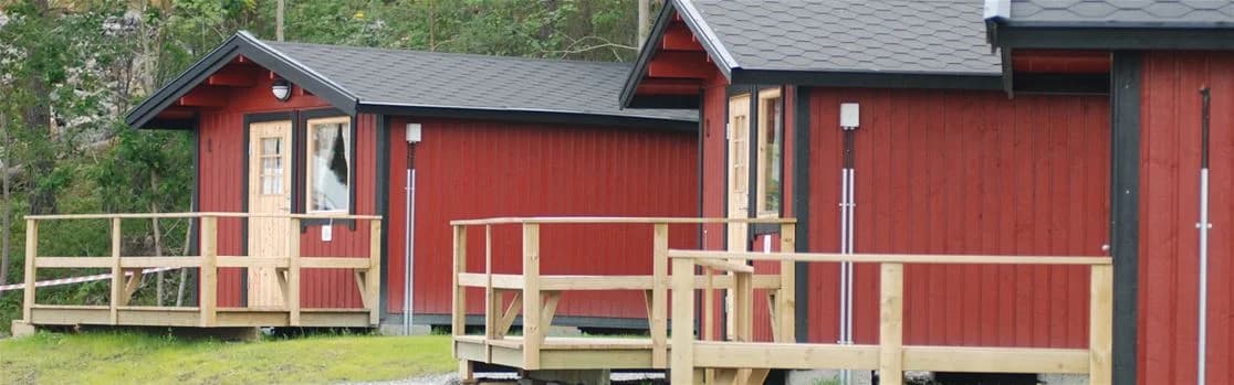 En rustik timmerstuga med träpaneler belägen i en grönskande miljö vid Arkösunds camping, omgiven av trädgårdsbyggnader.