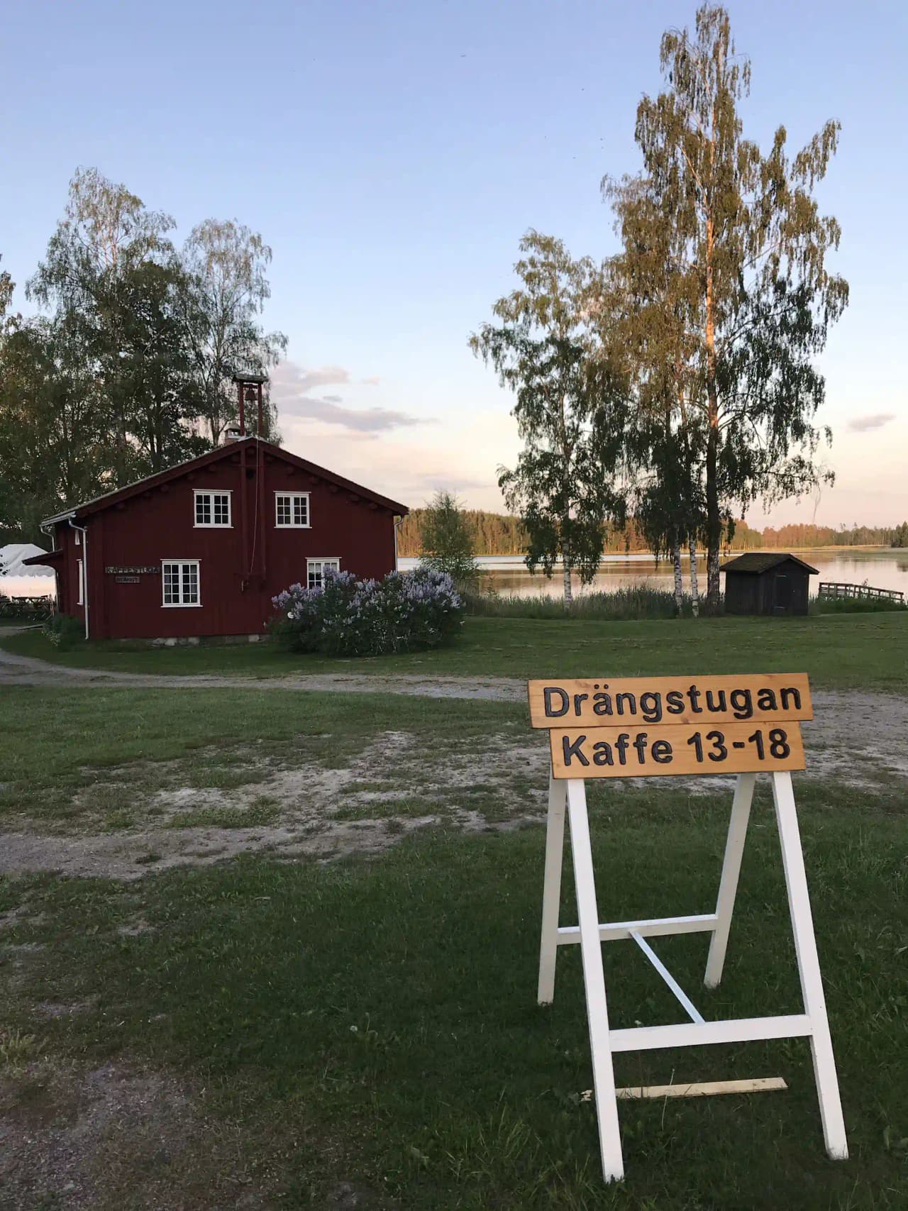 En grön gräsmatta i skuggan med ett utomhusbord och stolar, omgivna av träd och buskar på Brunskog hembygdsgård.
