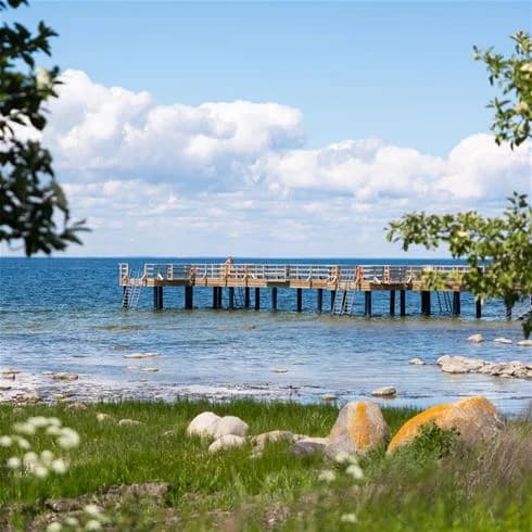En blå himmel med moln ovanför ett vidsträckt vattendrag. I förgrunden sträcker sig en brygga ut mot horisonten, omgiven av kustnära och oceaniska landformer.