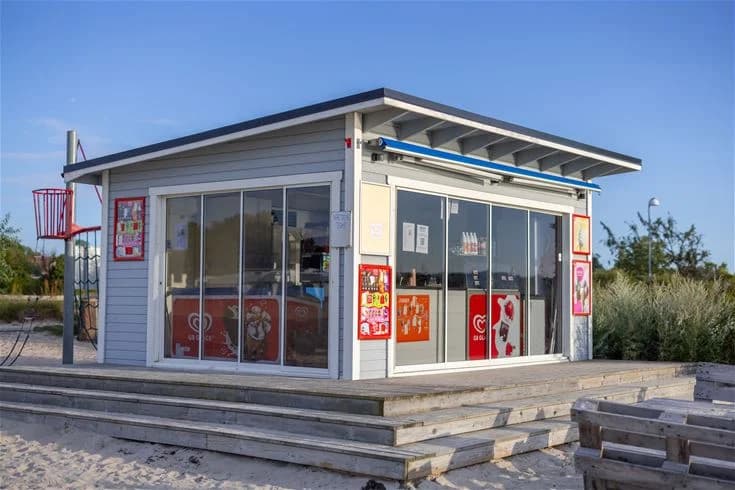 En campingplats med en liten kiosk under skugga, där det finns reklamskyltar och allmänna ytor för besökare.