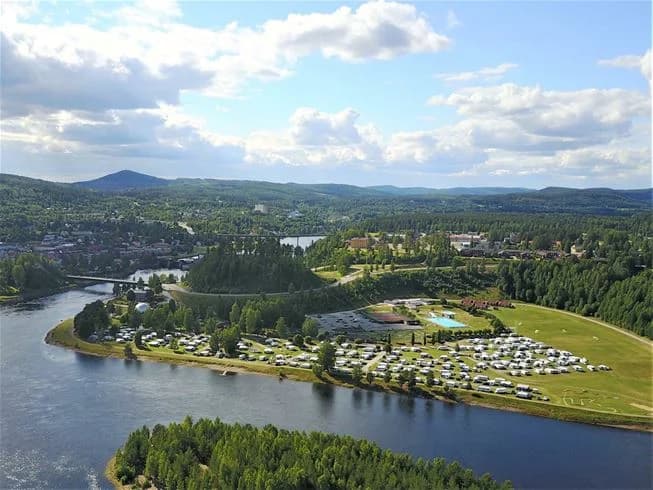 Flygfoto över Sollefteå Camping visar en campingplats vid vattnet, omgiven av en flod och grönskande landskap.