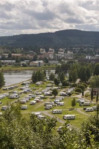 En campingplats vid en sjö med uppställda husbilar och husvagnar, omgiven av grönskande natur och berg.