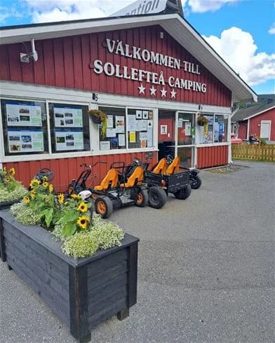 En logotyp för Sollefteå Camping, omgiven av grönska och reklam som visar campingens bekvämligheter och aktiviteter.