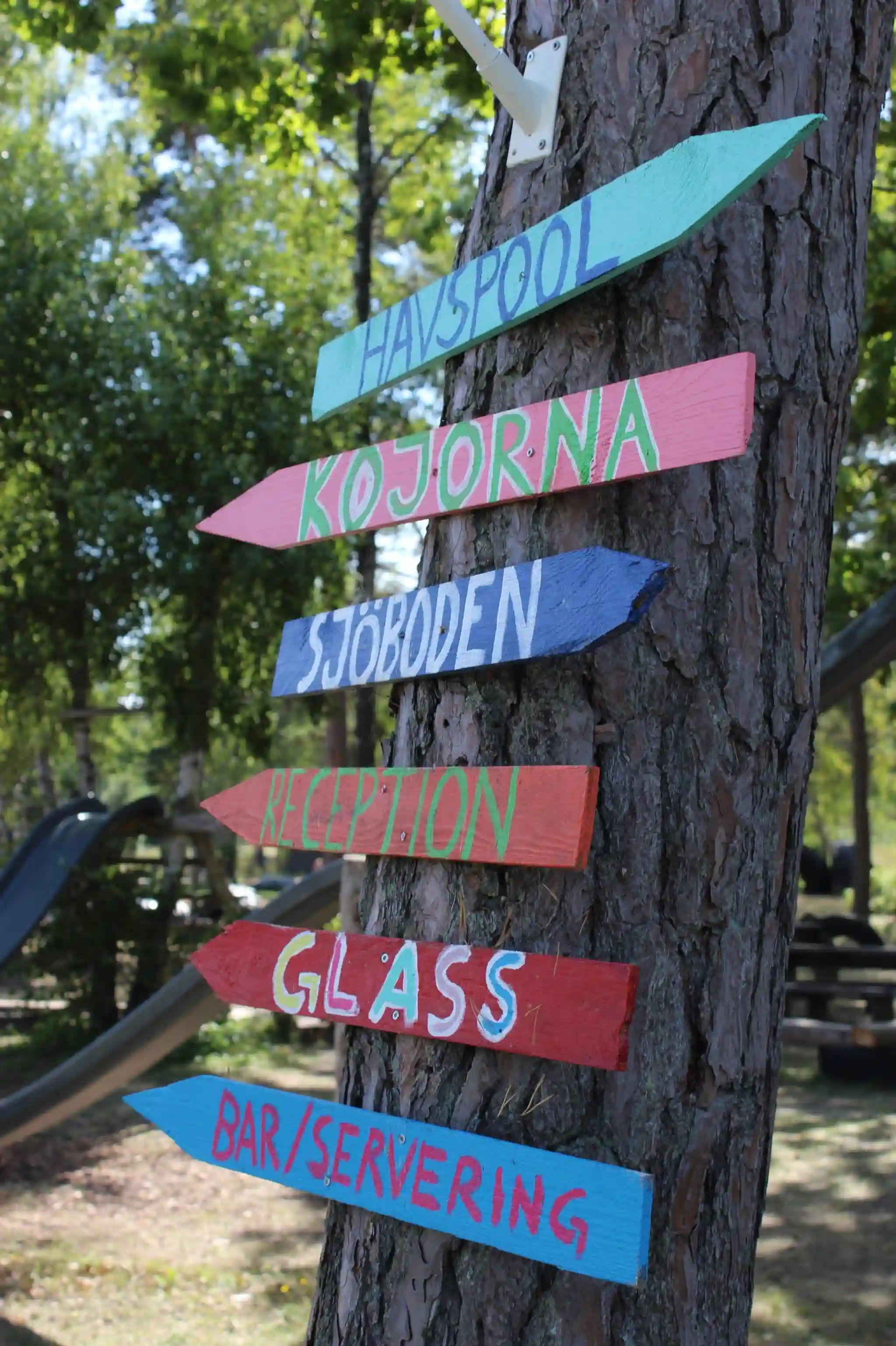 En träskylt vid infarten till Fide Äventyrsby & Camping, omgiven av grönska och träd.