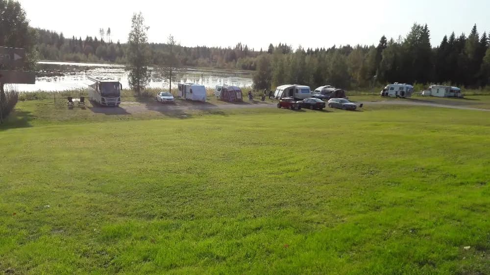Bild av Gammeltomtens camping: En öppen gräsmark intill en sjö med husbilar parkerade. Campingen omges av en lummig äng och är inramad av skogsklädda kullar i bakgrunden.