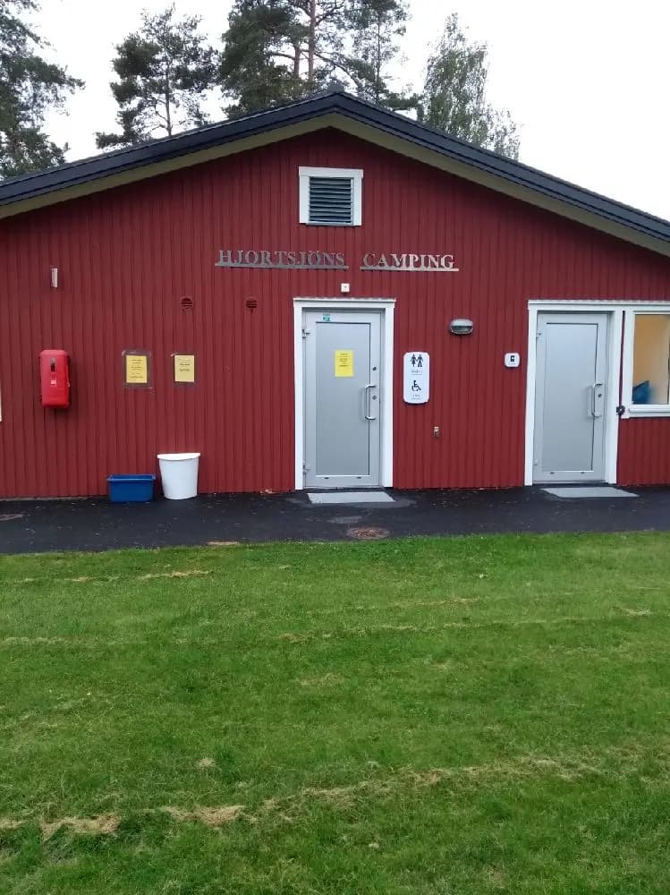 En liten byggnad med en dörr, omgiven av en gräsmatta med träpaneler på sidorna, vid Hjortsjöns camping.