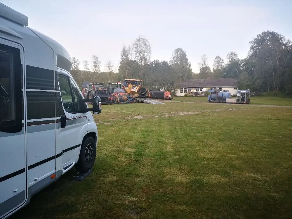 En grönskande hembygdsgård med en parkeringsplats där flera husbilar och skåpbilar står uppradade på en grön gräsmatta.