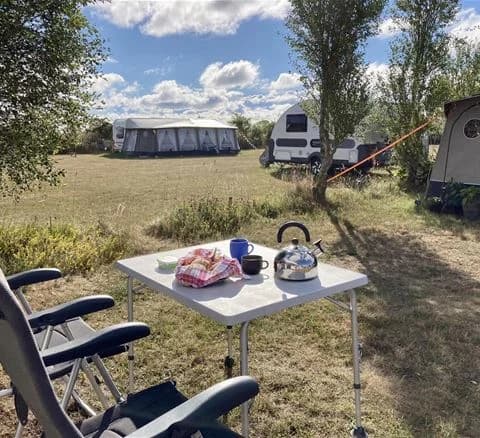 Bild av Wikegårds semesterby & ställplatscamping, med tält, husvagnar, en husbil, en skuggande presenning och utemöbler i en grönskande parkmiljö. En plats för avslappnad utomhusrekreation.