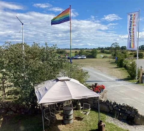 Bild av Wikegårds semesterby & ställplatscamping som visar en parkeringsplats med flera fordon, inklusive en familjebil och en SUV. Det finns flaggor som vajar på stolpar och en tältliknande konstruktion i bakgrunden.