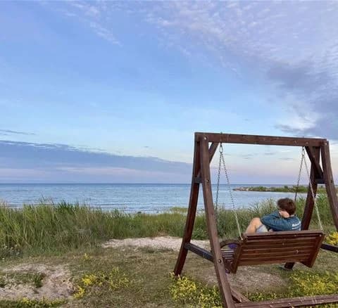 En somrig kustvy vid Wikegårds Semesterby & Ställplatscamping, med blå himmel, moln, och ett glittrande hav i bakgrunden. På bilden syns även utemöbler på en grönskande plats nära kusten.