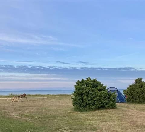 En campingplats med ett tält uppställt på en grön äng under en klarblå himmel med några moln. I bakgrunden syns ett öppet landskap, vilket skapar en fridfull och naturnära atmosfär.