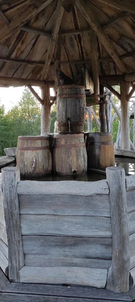 En träbyggnad omgiven av naturliga material, med ett traditionellt brunnslock och träplankor i förgrunden, på Lufta Camping & Restaurang.