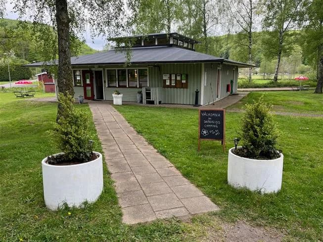 En grön gräsmatta med en liten röd stuga i bakgrunden, omgiven av träd och buskar i en lummig trädgårdsmiljö. Ett utomhusbord i skuggan står på gräset, idealiskt för avkoppling i parkliknande omgivningar.