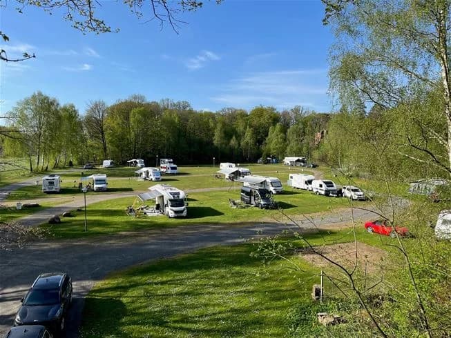 En campingplats i lantlig miljö med träd i bakgrunden. Det finns flera husbilar, husvagnar och parkerade bilar.