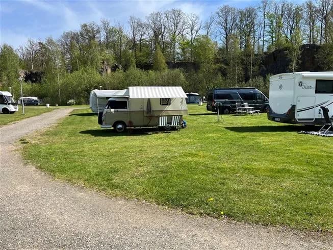 En husvagn står uppställd på en grön gräsmatta vid Skäralids camping, omgiven av träd och parkerade bilar på en solig dag.