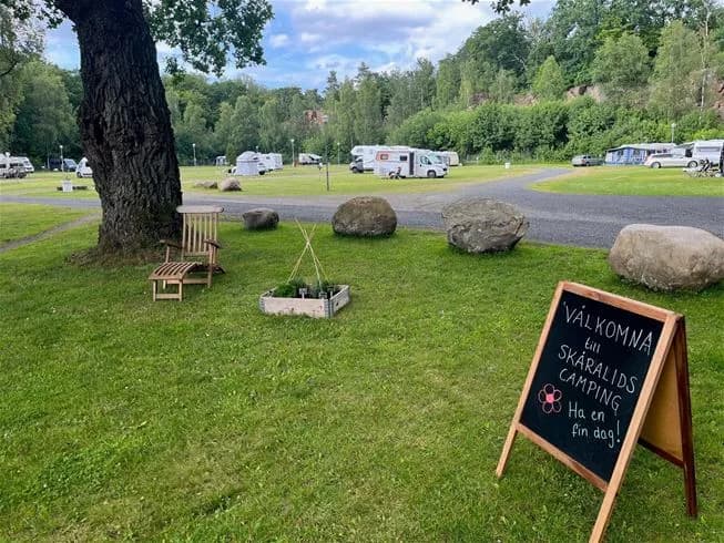 En grönskande campingplats med träd och gräs. Det finns picknickbord och en husbil i området, vilket ger en inbjudande plats för rekreation och avkoppling.