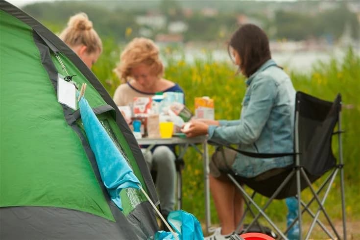 En sommardag på Lisebergs camping i Askim Strand där människor njuter av friluftsliv. Flera tält är uppslagna på området och besökare sitter bekvämt runt omkring och deltar i olika fritidsaktiviteter.
