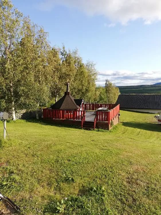 En grönskande trädgård med välklippt gräsmatta i skugga, inramad av staket och träd. Det finns picknickbord och en enkel uteplats. I bakgrunden syns ett litet trädgårdsskjul.