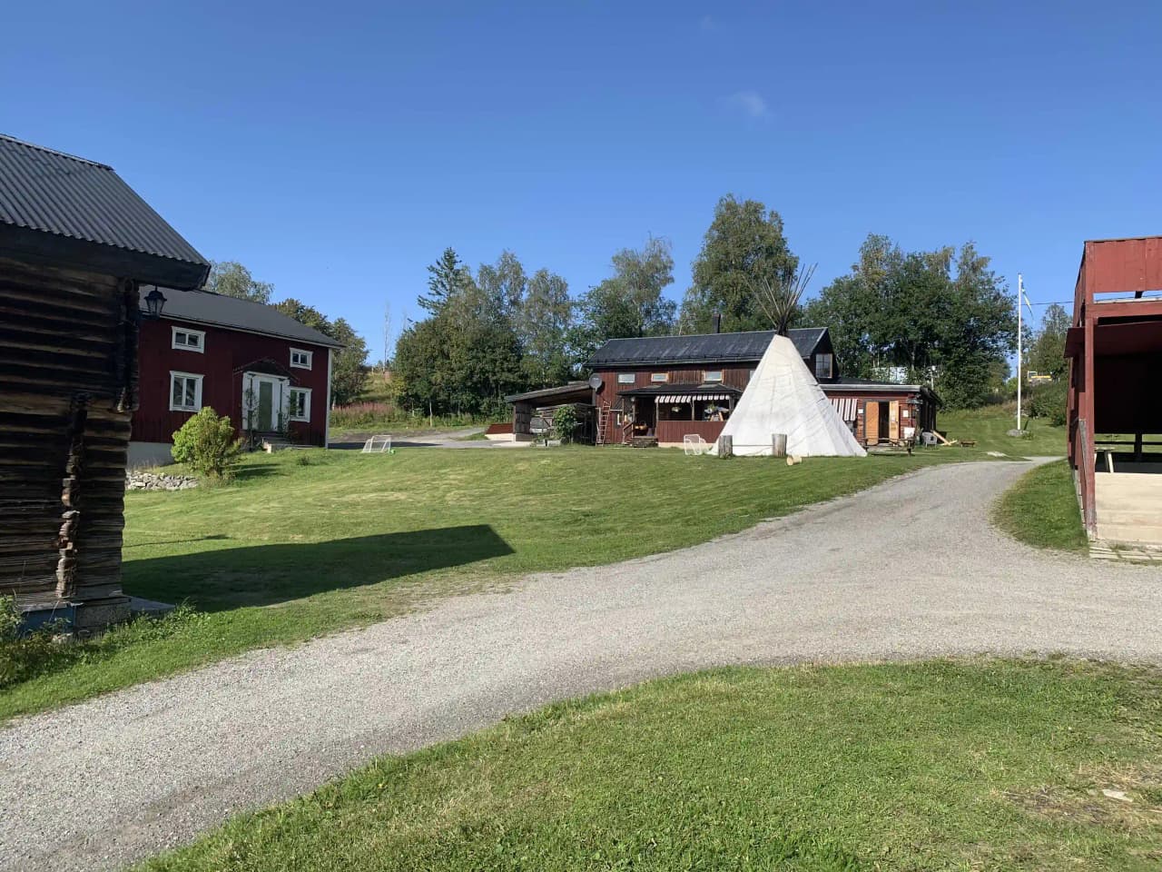 En grön äng sträcker sig framför en enkel stuga på Kvarnsjö Camp, omgiven av lantlig natur och en liten by i bakgrunden.