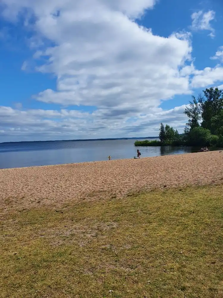 En vidsträckt himmel med lätta moln sträcker sig över en somrig strand vid Hjälmargården & Läppe Camping. Horisonten smälter samman med den blåa himlen och det kustnära landskapet skapar en inbjudande atmosfär för besökare.