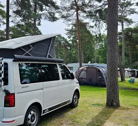 En äldre Volkswagen-buss, parkerad på en campingplats vid Ystad, med registreringsskylt och detaljerade delar som dörrar och stötfångare synliga. I bakgrunden skymtar campingmiljön.