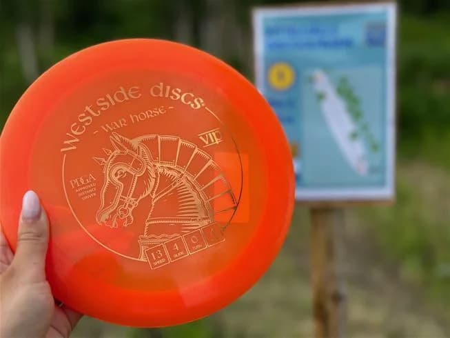En person kastar en frisbee på en grön gräsplan vid Camp Kittelfjäll. I bakgrunden syns skog och berg, vilket skapar en naturskön miljö.