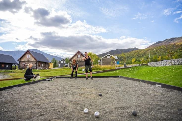 En naturskön vy över Camp Kittelfjäll med frodiga gröna kullar och dalar. I bakgrunden syns mäktiga fjälltoppar som sträcker sig mot himlen. I förgrunden finns en stuga belägen på en grässluttning, inbjudande för besökare att koppla av och njuta av den vackra omgivningen.
