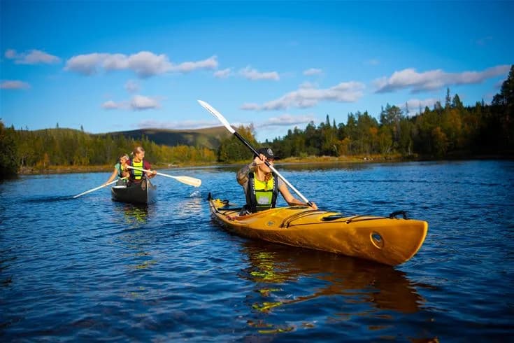 En grupp personer paddlar kajak på en lugn sjö omgiven av berg och skog vid camp Kittelfjäll.