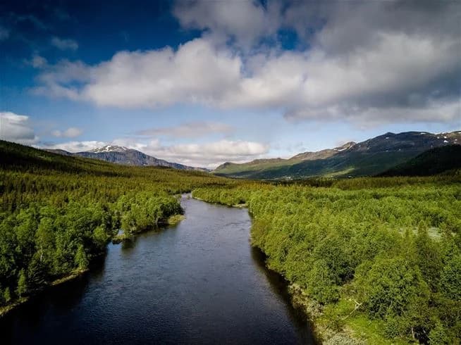 En pittoresk vy över Kittelfjäll med majestätiska berg och en klar vattenyta i förgrunden, omgiven av grönska och moln på himlen.