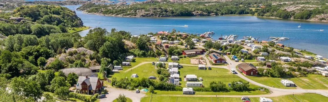 En fågelperspektivvy över Rörviks camping vid en kusten, med en vik och små bybyggnader omgivna av frodig natur och glittrande vatten.
